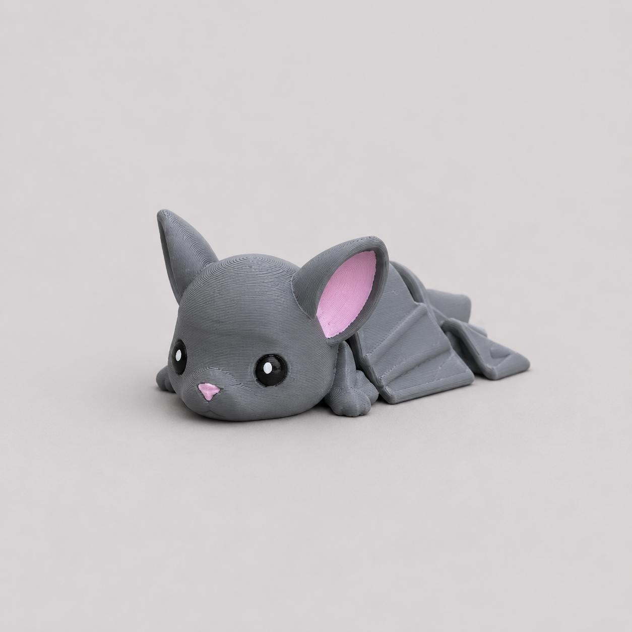 3D printed Teeny Bat figure in cool grey with pink inner ears and outstretched wings, lying down